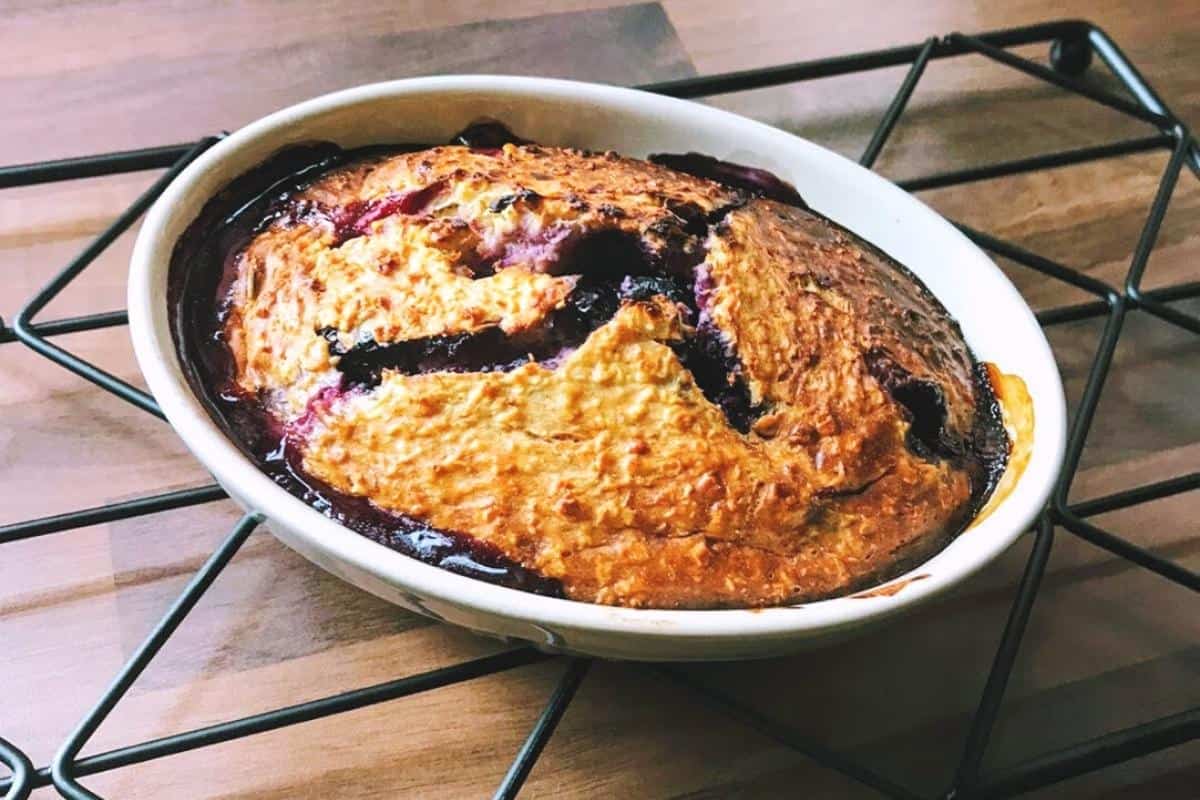 A freshly baked berry cobbler, reminiscent of Slimming World baked oats, sits in an oval dish on a metal cooling rack. It has a golden-brown crust with purple berry filling peeking through cracks, all set against a wooden countertop.