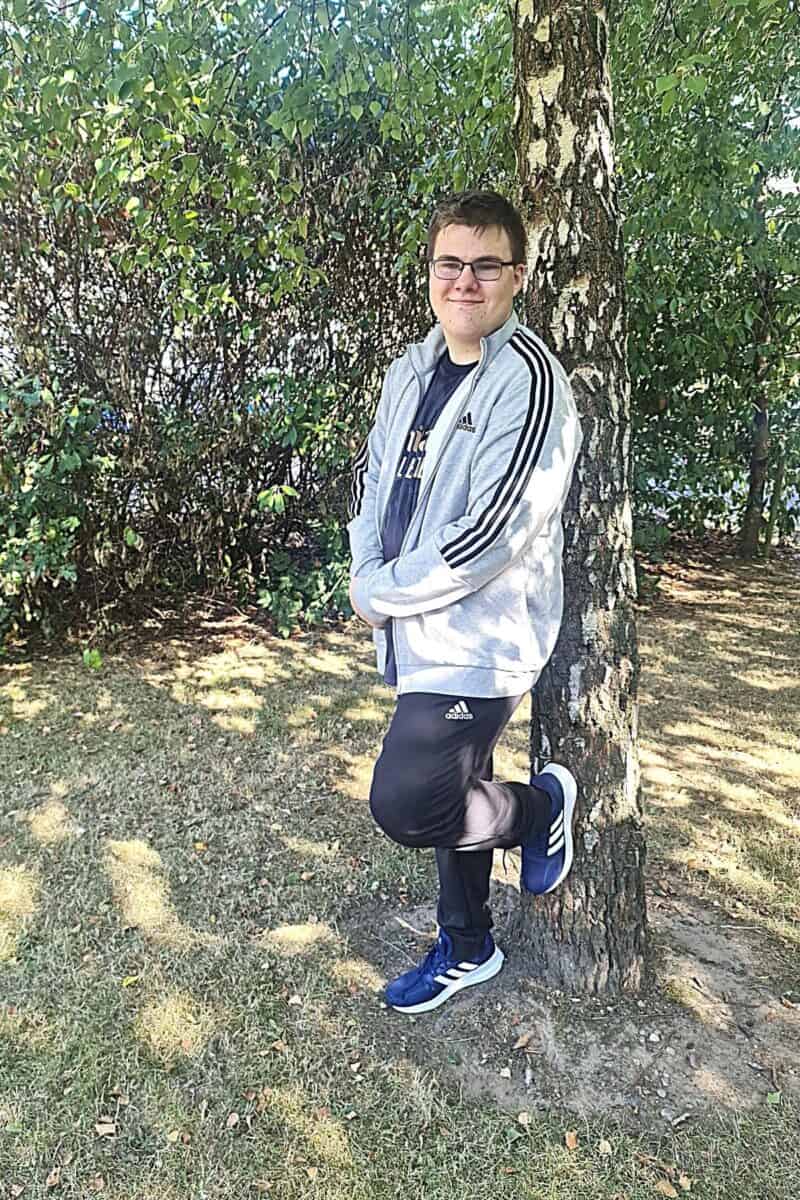 A person in glasses leans against a tree, wearing a gray jacket, dark pants, and blue shoes. Standing on grassy ground with bushes and sunlit leaves in the background, they take a moment to reflect before starting college.
