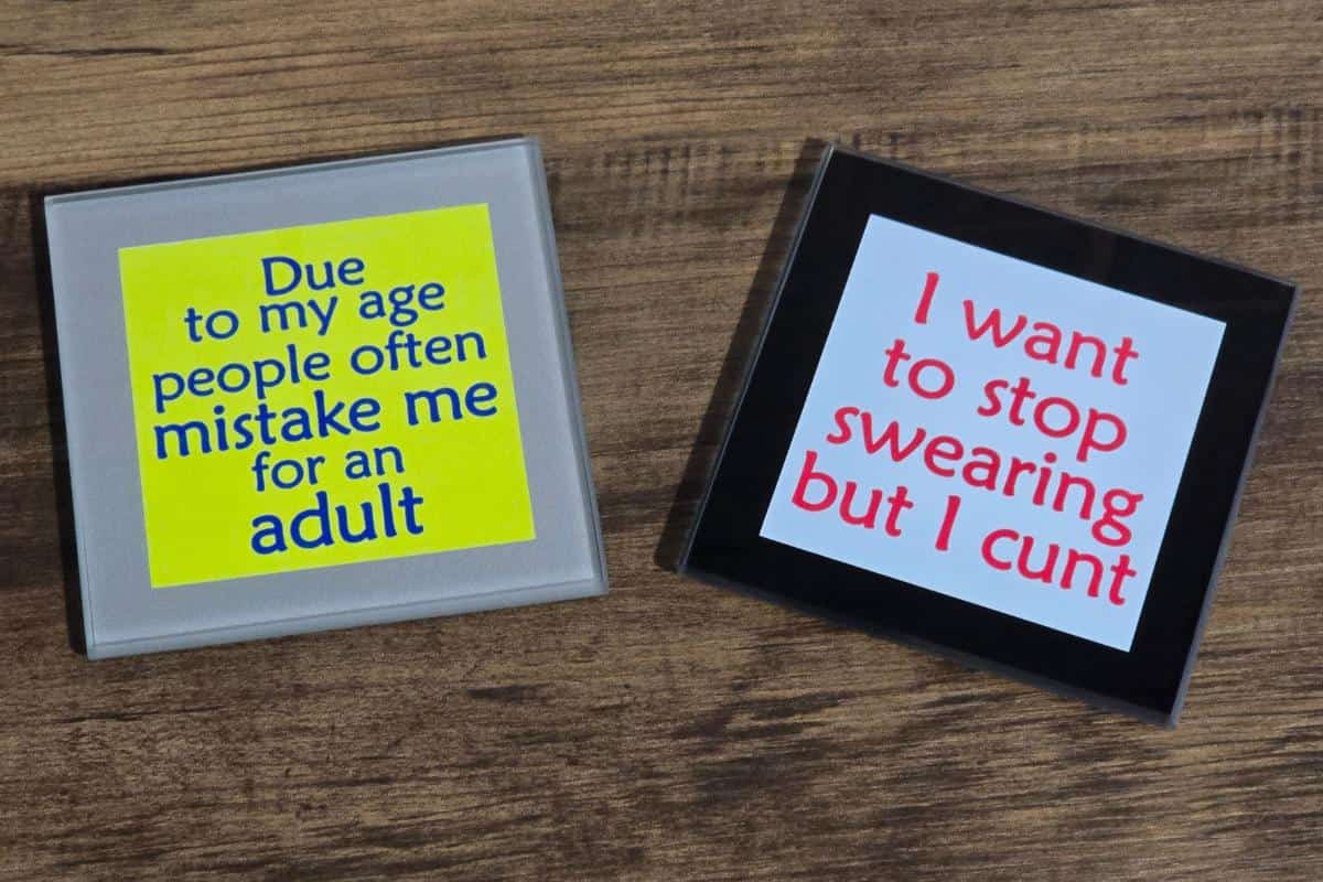 Two square coasters from the Rude Gift Website rest on a wooden surface. One reads, "Due to my age people often mistake me for an adult," while the other quips, "I want to stop swearing but I cunt." Perfect for those who appreciate a Mr. Inappropriate Review-worthy chuckle.