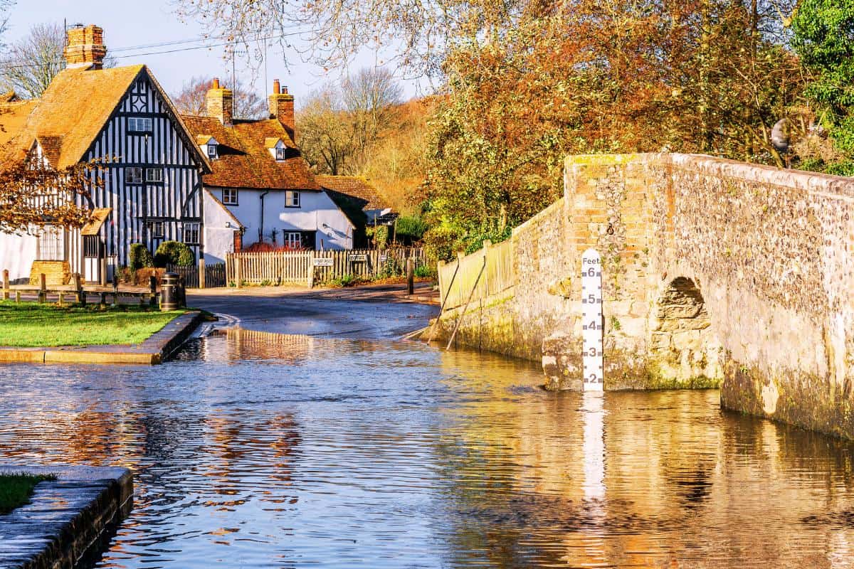A scenic village scene with a half-timbered house next to a narrow road. A stone bridge crosses a shallow river, marked with a water depth gauge. Trees with autumn foliage line the background.