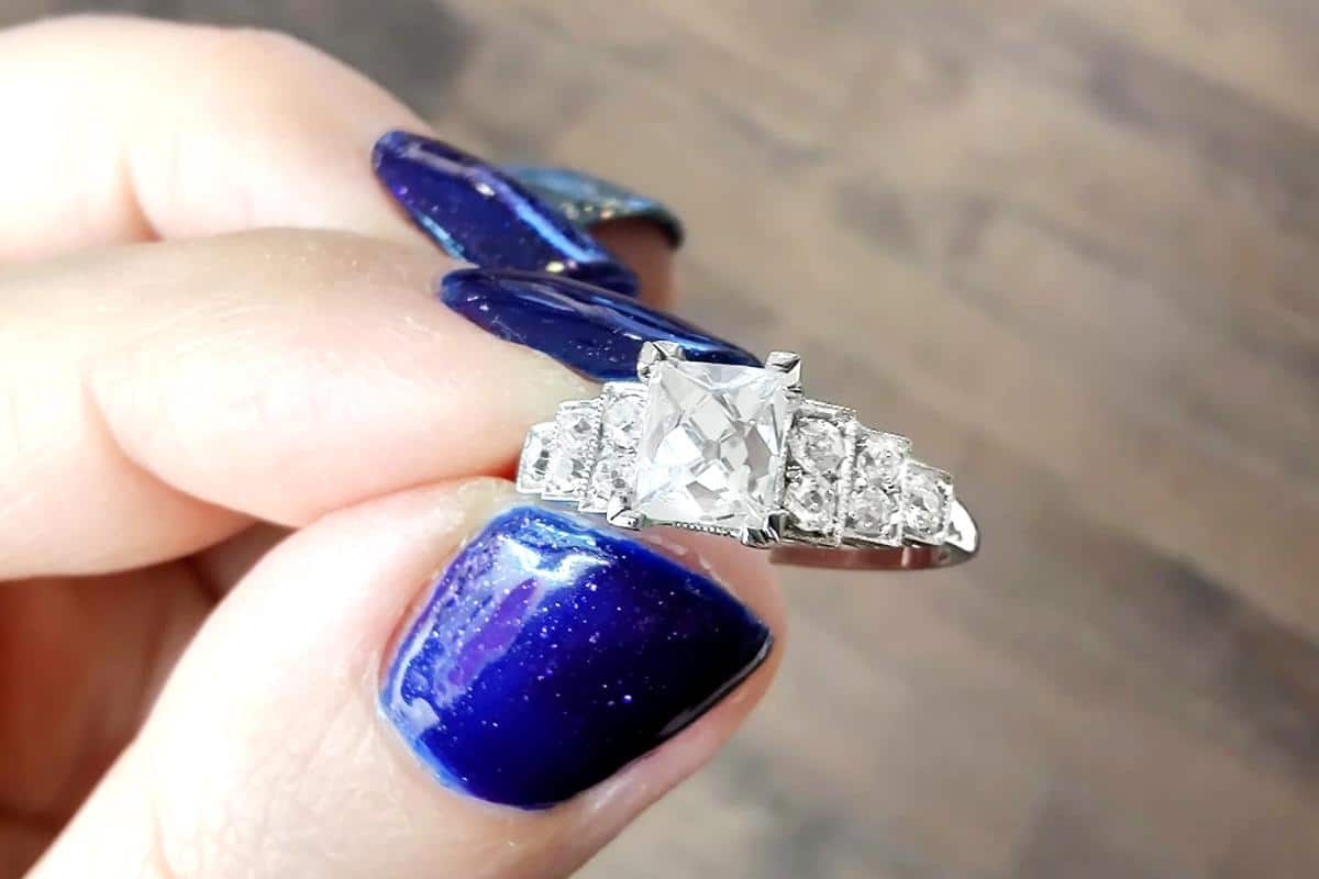 A person with blue sparkly nail polish holds a silver ring featuring a large rectangular clear gemstone in the center, flanked by smaller round clear stones on each side. The background is a blurred wooden surface.