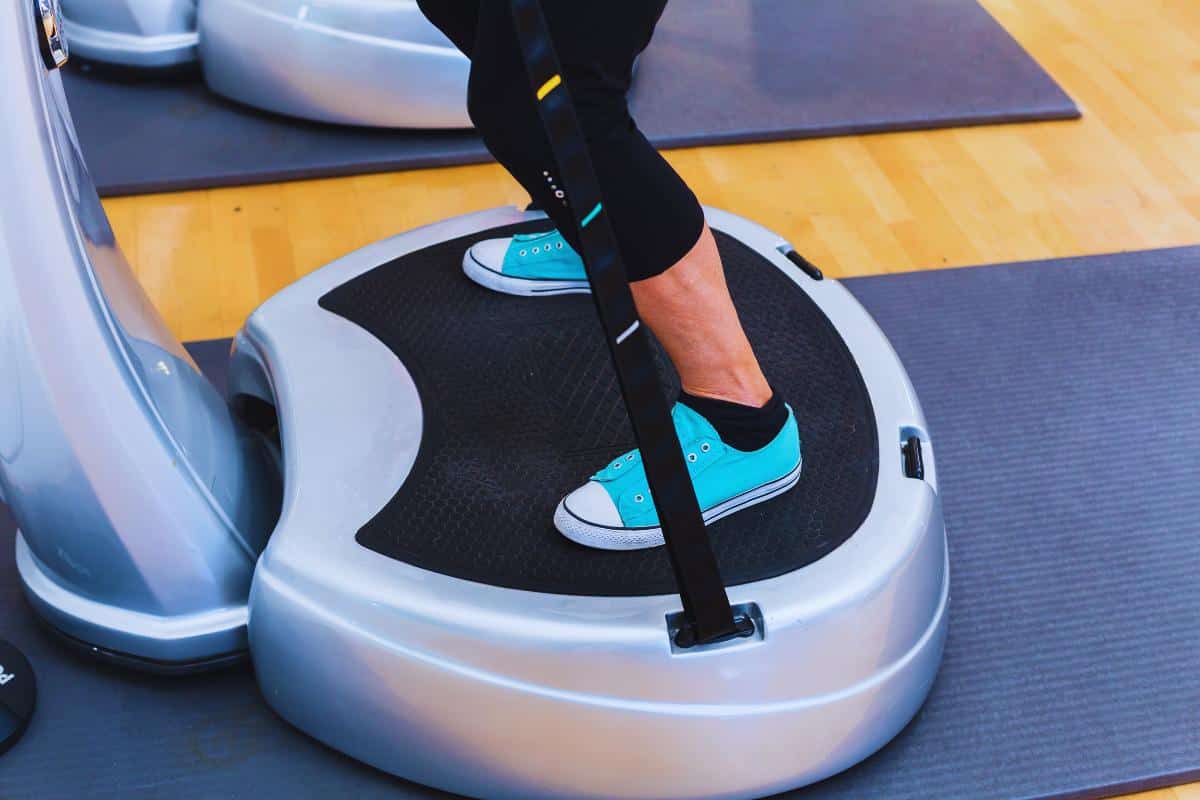 Standing on the vibration plate, a person in teal sneakers and black leggings engages in their weight loss journey. The wooden floor beneath them, with exercise mats nearby, sets the stage for understanding how to use this innovative fitness tool effectively.