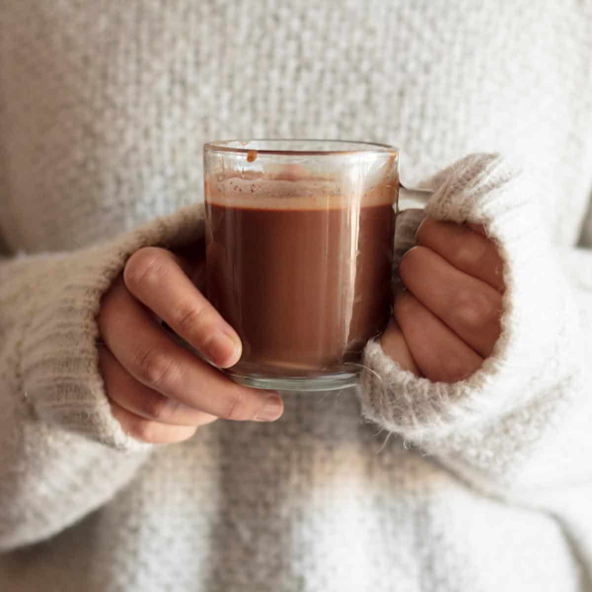 ladies hands with cream jumper in background holding a glass mug of hot chocolate made in a chocolate maker