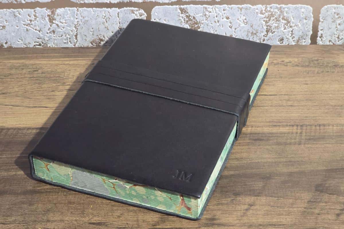 Black leather-bound book with embossed initials "JM," lying closed on a wooden surface, framed by a brick wall in the background—an elegant choice for any Mother's Day gift guide.