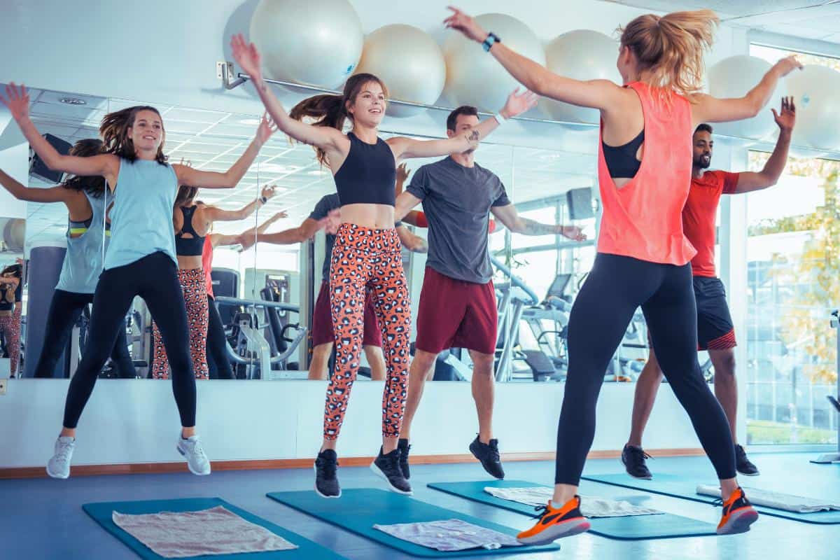 People are engaged in a group exercise class at a gym, executing jumps on mats. Clad in athletic wear, they make the most of their busy schedules. Large exercise balls and gym equipment reflect off a large mirror, showcasing an intense yet efficient 15-minute workout session.