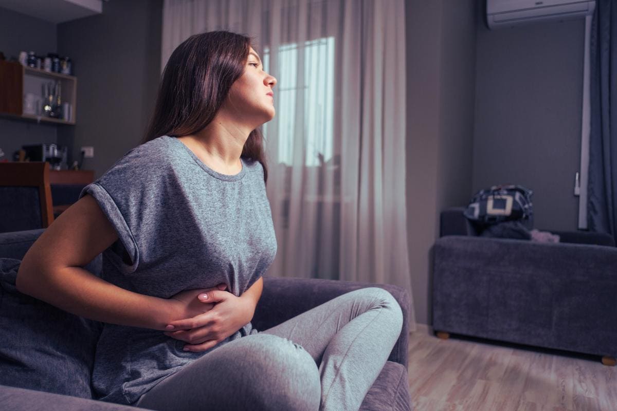 A woman sits on a gray sofa in the living room, wearing a gray t-shirt and leggings. Pressing her hands against her abdomen, she seems to be in discomfort, possibly from gallstones. The room is furnished simply with curtains, a table, and a chair.