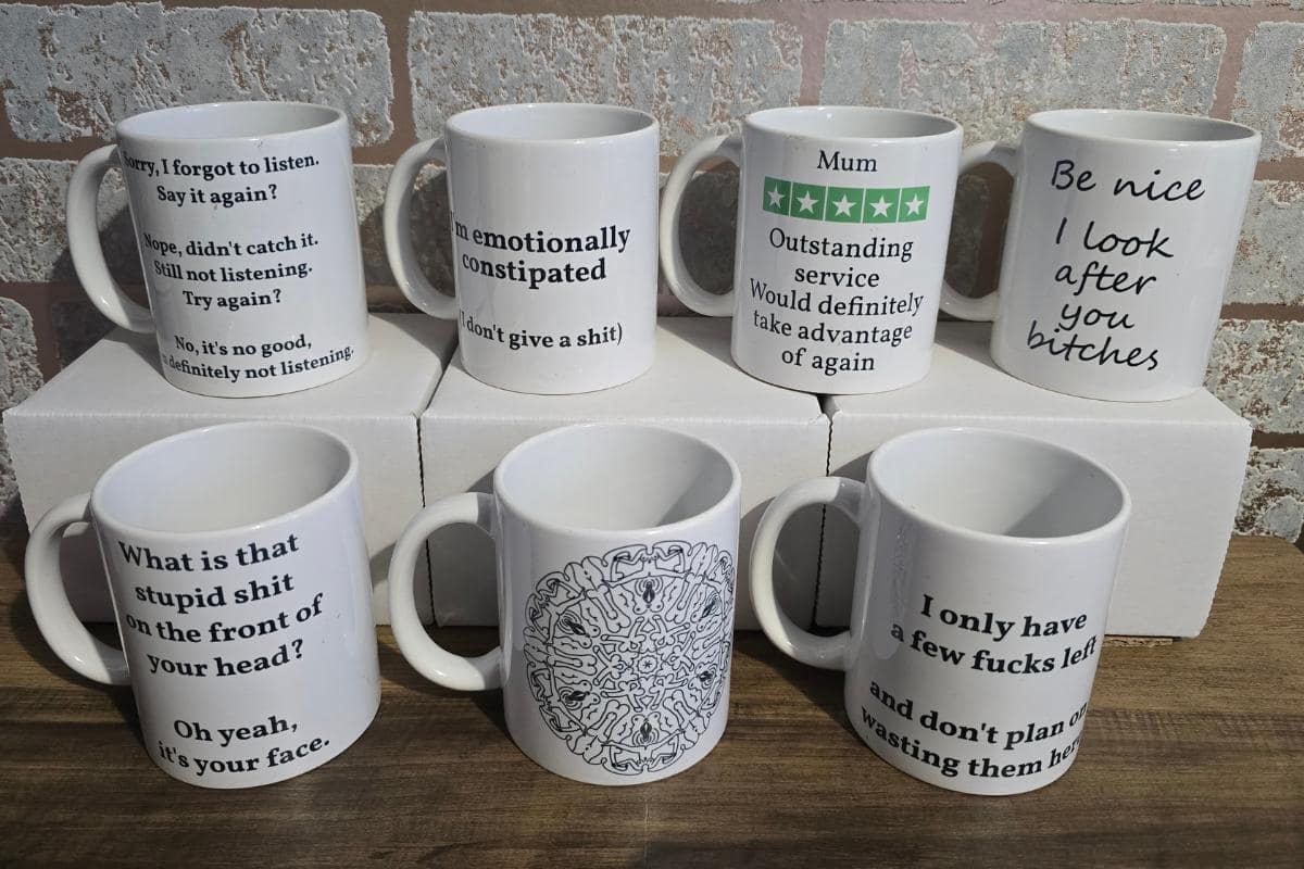 Seven white mugs featuring humorous and sarcastic text phrases are displayed on a wooden surface and white boxes, with a brick wall in the background. Perfect for those who enjoy a chuckle or two, they make ideal finds on the Laugh and Blush section of a rude gift website.