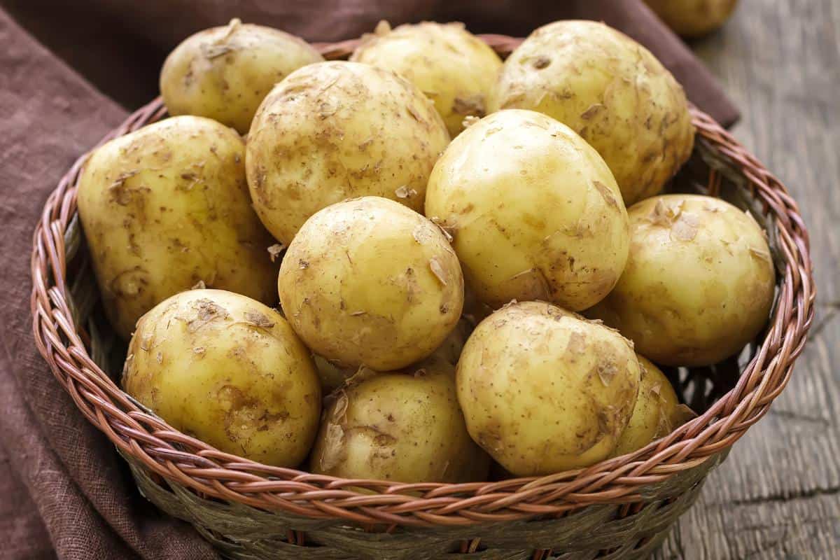 A woven basket filled with several unpeeled potatoes rests on a wooden surface. The fresh spuds, dusted with dirt, await their fate—perhaps even to freeze. A brown cloth is partially visible in the background, adding a rustic touch to the scene.