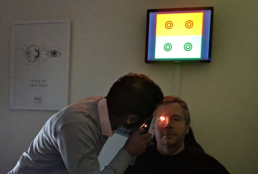 Eye examination in progress at Marks and Spencer Opticians. A healthcare professional uses an ophthalmoscope to check a patient's eyes, while a screen displays colorful test patterns in the background.