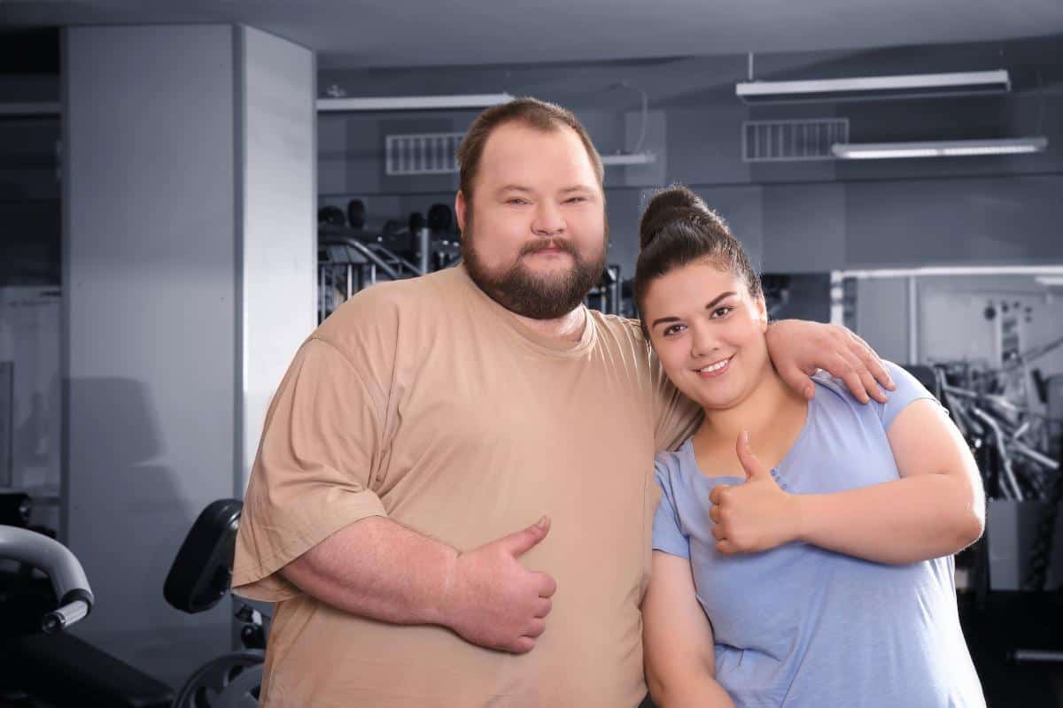 Couple of overweight people with their thumbs up in a gym.