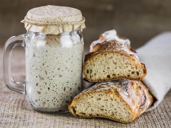 A jar of bubbling sourdough starter sits next to two sliced pieces of rustic bread on a burlap surface.
