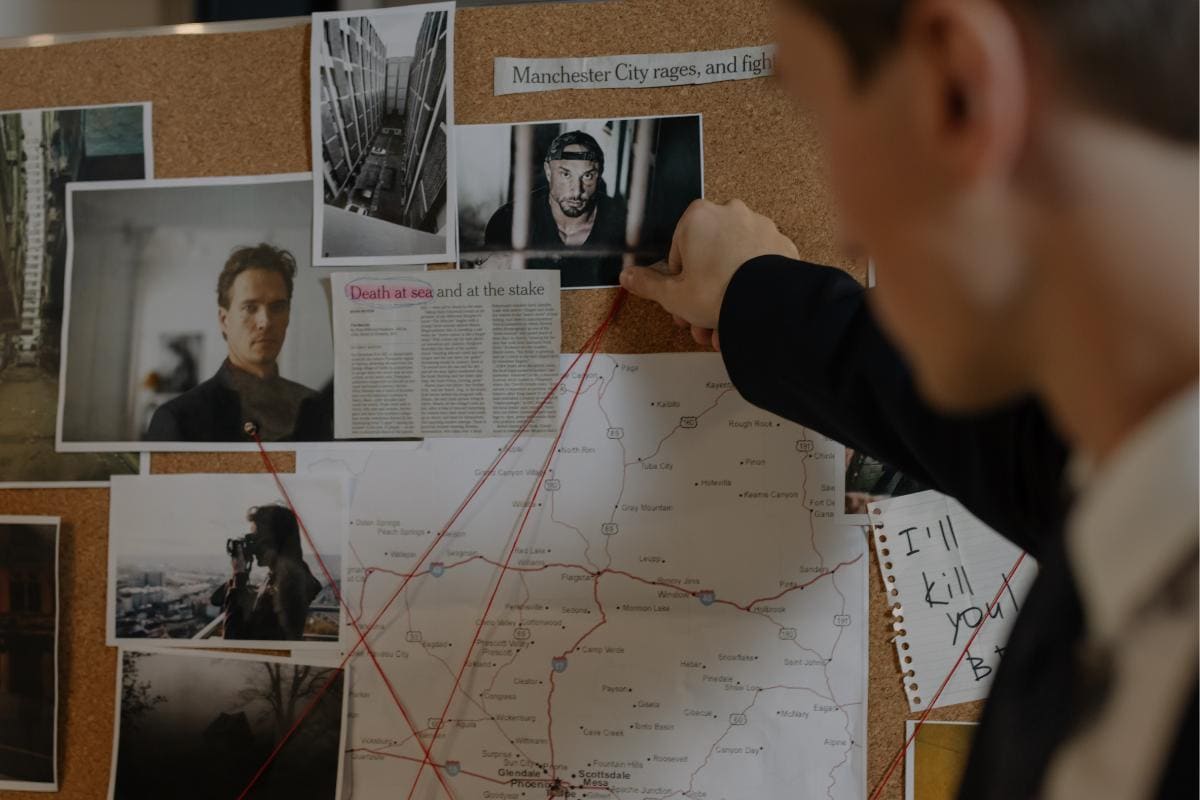 A person in a suit points to a photo on a bulletin board covered with various images, papers, and a map. Some text reads "Death at sea and at the stake." Like something out of a true crime podcast, red strings connect different points on the board.